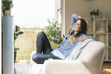Happy woman with a glass of infused water sitting on the couch at home - UUF17230