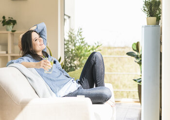 Happy woman with a glass of infused water sitting on the couch at home - UUF17231