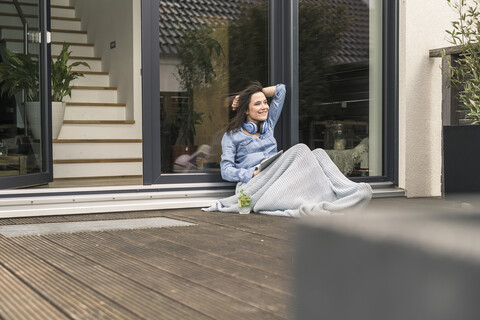 Glückliche Frau, eingewickelt in eine Decke, sitzt auf der Terrasse zu Hause mit Tablet, lizenzfreies Stockfoto