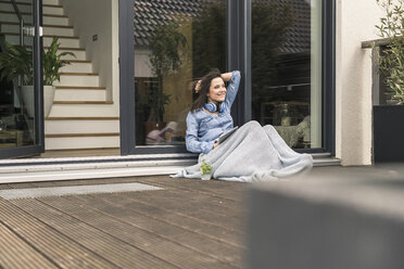 Glückliche Frau, eingewickelt in eine Decke, sitzt auf der Terrasse zu Hause mit Tablet - UUF17244