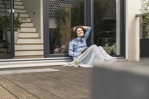 Glückliche Frau, eingewickelt in eine Decke, sitzt auf der Terrasse zu Hause mit Tablet, lizenzfreies Stockfoto