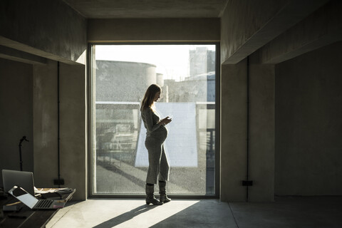 Schwangere Geschäftsfrau, die am Bürofenster steht und ein Smartphone benutzt, lizenzfreies Stockfoto