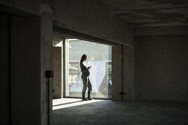 Schwangere Geschäftsfrau, die am Bürofenster steht und ein Smartphone benutzt - MJRF00185
