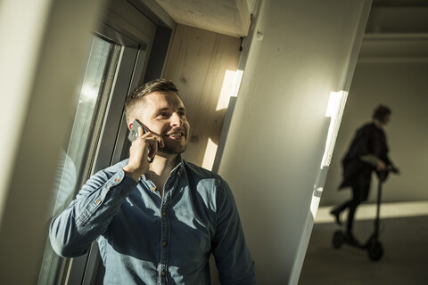 Junger Geschäftsmann beim Telefonieren, während ein Kollege im Hintergrund ein Kickboard benutzt, lizenzfreies Stockfoto
