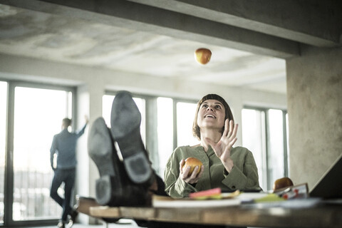 Frau jongliert mit Äpfeln im Büro, sitzt mit den Füßen auf dem Schreibtisch, lizenzfreies Stockfoto