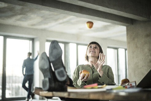 Woman juggling apples in the office, sitting with feet on desk - MJRF00204