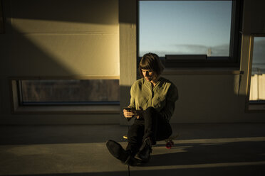 Businesswoman sitting on office floor at sunset, using smartphone - MJRF00226