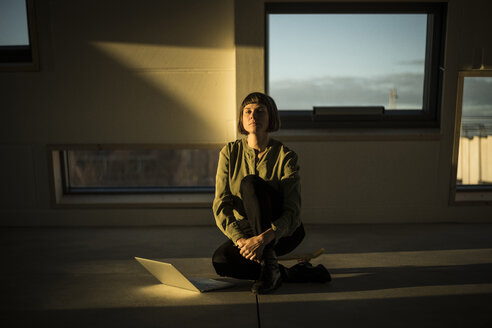 Geschäftsfrau mit Laptop, sitzend auf dem Büroboden bei Sonnenuntergang, mit geschlossenen Augen - MJRF00230