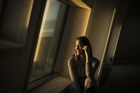 Frau sitzt am Bürofenster bei Sonnenuntergang und telefoniert, lizenzfreies Stockfoto