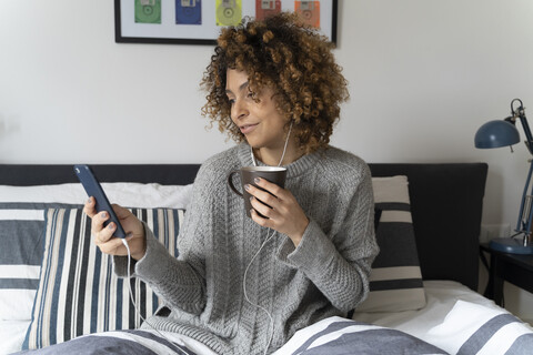 Frau sitzt auf dem Bett, trinkt Kaffee, benutzt Smartphone und Kopfhörer, lizenzfreies Stockfoto