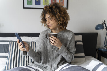 Frau sitzt auf dem Bett, trinkt Kaffee, benutzt Smartphone und Kopfhörer - FMOF00538