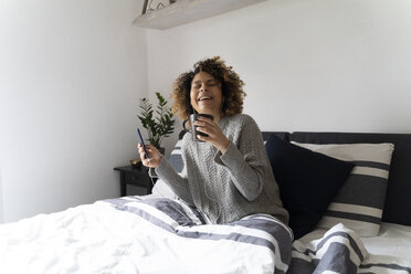 Woman sitting on bed, drinking coffee, using smartphone and earphones - FMOF00539