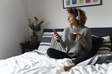 Frau sitzt auf dem Bett, trinkt Kaffee, benutzt Smartphone und Kopfhörer - FMOF00540