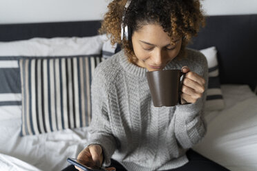 Frau sitzt auf dem Bett, trinkt Kaffee und hört Musik mit Kopfhörern und Smartphone - FMOF00542