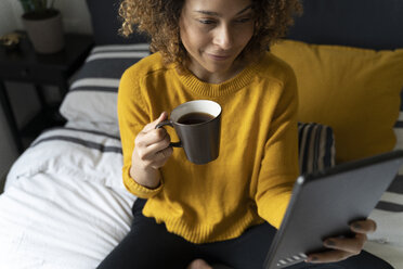 Frau auf dem Bett sitzend, eine Pause machend, Kaffee trinkend, auf ein digitales Tablet tretend - FMOF00570