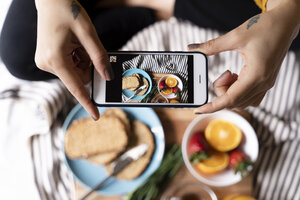 Frau auf dem Bett sitzend, die mit ihrem Smartphone Fotos von ihrem Frühstück macht - FMOF00573