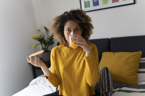 Frau sitzt auf dem Bett, trinkt Orangensaft, hält Toastbrot, lizenzfreies Stockfoto