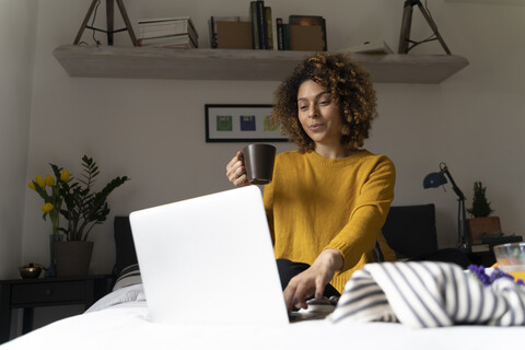 Frau sitzt auf dem Bett, benutzt einen Laptop und trinkt Kaffee, lizenzfreies Stockfoto