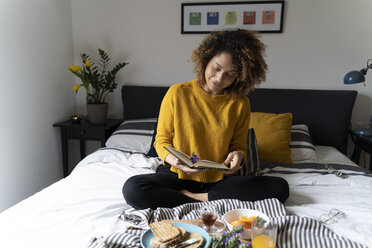 Frau sitzt auf dem Bett, frühstückt gesund und liest ein Buch - FMOF00578
