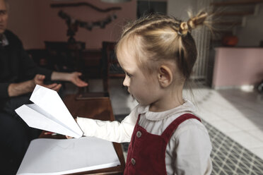 Mädchen hält Papierflugzeug im Wohnzimmer mit Großvater im Hintergrund - KMKF00820