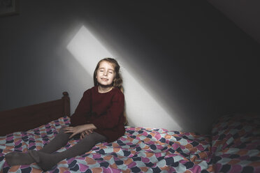 Smiling girl sitting on bed at home with closed eyes - KMKF00840