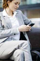 Junge Geschäftsfrau, die auf einer Bank in der Stadt sitzt und ein Smartphone benutzt - JRFF03051