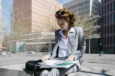 Junge Geschäftsfrau, die an einem sonnigen Tag auf einer Bank in der Stadt sitzt und Dokumente liest - JRFF03069