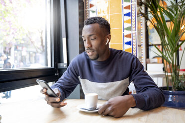Junger Mann mit drahtlosen Kopfhörern in einem Café, der auf sein Handy schaut - OCMF00399