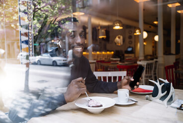 Portrait of smiling young man with earphones and smartphone behind windowpane in a restaurant - OCMF00401