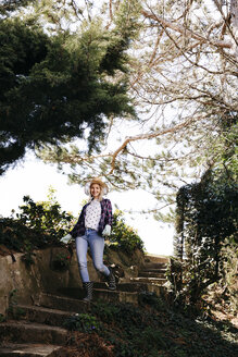 Junge blonde Frau mit Strohhut geht eine Gartentreppe hinunter - HMEF00335