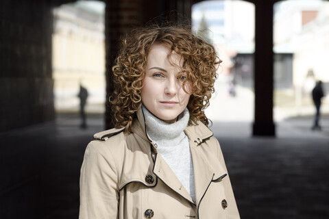 Porträt einer Frau mit lockigem Haar, die einen beigen Trenchcoat und einen Rollkragenpullover trägt, lizenzfreies Stockfoto