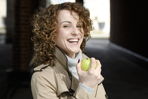Porträt einer lachenden Frau mit lockigem Haar und grünem Apfel, lizenzfreies Stockfoto