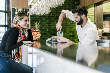 Lächelnde Frau am Tresen einer Bar, die den Barkeeper bei der Zubereitung eines Cocktails beobachtet - ERRF01126