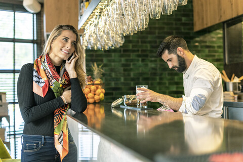 Lächelnde Frau am Handy am Tresen einer Bar mit Barkeeper bei der Zubereitung eines Cocktails, lizenzfreies Stockfoto