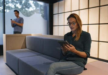 Junge Frau sitzt auf einer Couch und benutzt ein Tablet mit einem Mann im Hintergrund - GUSF01888