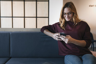 Lächelnde junge Frau sitzt auf der Couch und benutzt ein Mobiltelefon - GUSF01928