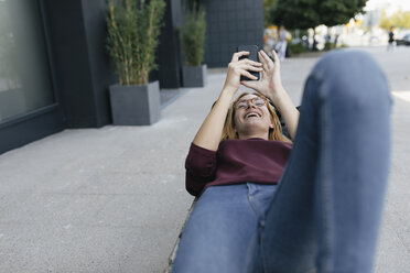 Glückliche junge Frau liegt auf einer Tasche und benutzt ein Mobiltelefon - GUSF01941