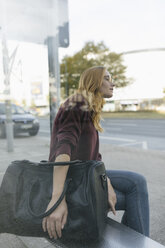 Junge Frau sitzt mit Tasche an der Bushaltestelle - GUSF01943
