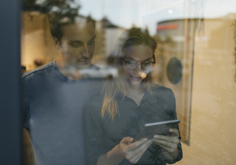 Junger Mann und Frau teilen sich ein Tablet hinter einer Fensterscheibe, lizenzfreies Stockfoto