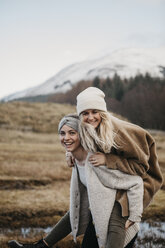 UK, Scotland, happy young woman carrying friend piggyback in rural landscape - LHPF00546