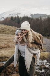 UK, Scotland, happy young woman carrying friend piggyback in rural landscape - LHPF00547