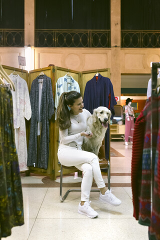 Lächelnde Frau mit Hund sitzt auf einem Stuhl in einer Vintage-Boutique, lizenzfreies Stockfoto