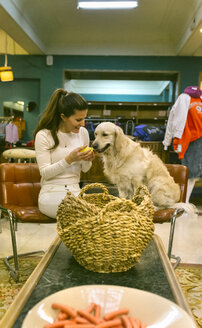 Lächelnde Frau mit Hund sitzt auf einem Stuhl in einer Vintage-Boutique - MGOF03999