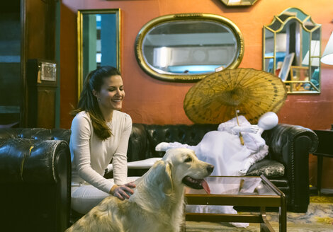Lächelnde Frau mit Hund, die auf einer Couch in einem Vintage-Laden sitzt - MGOF04007