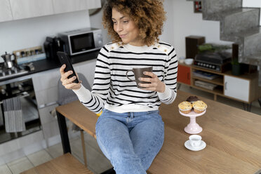 Woman sitting at home, using smartphone, eating muffin - FMOF00590