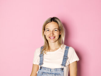 Porträt einer lächelnden blonden Frau in Latzhose vor einem rosa Hintergrund - HMEF00339