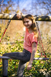 Porträt eines lächelnden Mädchens, das auf einer Gymnastikstange im Garten sitzt - SARF04234