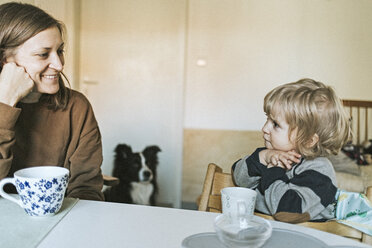 Lächelnde Mutter und Tochter sitzen am Tisch zu Hause mit Hund im Hintergrund - DWF00419