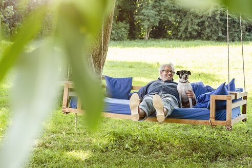 Ein älterer Mann entspannt sich mit seinem Hund auf einem Schaukelbett in seinem Garten - PESF01616