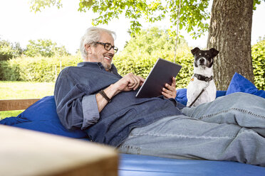 Ein älterer Mann entspannt sich auf einem Schaukelbett in seinem Garten und benutzt ein digitales Tablet - PESF01618